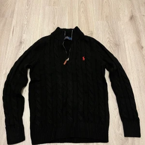 Svart stickad tröja Polo Ralph Lauren - Svart kabelstickad tröja från Polo Ralph Lauren med röd broderad logga på bröstet. Tröjan har lång ärm, ribbad krage och muddar samt en snygg half zip-dragkedja med läderdetalj. Perfekt för dig som gillar klassisk och stilren look.