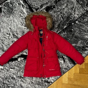 jacka  från Bondelid - Riktigt snygg röd pufferjacka från Bondelid med huva och fuskpäls. Jackan har flera fickor, dragkedja och knappar framtill, samt coolt Bondelid-märke på insidan. Fodret är mörkblått med gula detaljer. Perfekt för kalla dagar!