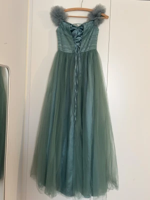 Grön långklänning med tyllaxlar - Drömmig grön långklänning med voluminösa tyllaxlar och omlottliv. Klänningen har ett svepande skirt tyg och snörning i ryggen för extra snygg passform. Perfekt för bal eller student! Materialet är lätt och ger en elegant känsla.