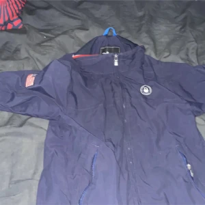 Marinblå vindjacka från Sail Racing - Snygg marinblå vindjacka från Sail Racing med dragkedja framtill och två sidofickor. Jackan har lång ärm, coolt Sail Racing-märke på bröstet och en patch med amerikansk flagga på ena ärmen. Perfekt för dig som gillar sportig och stilren look.