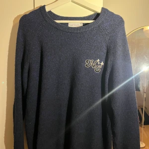 Marinblå stickad tröja från Naked Collection - Helt ny och Mysig marinblå stickad tröja från Naked Collection med broderad logga 'NC' och stjärnor på bröstet samt text på ryggen. Rund halsringning och långa ärmar, perfekt för att hålla dig varm under kyliga dagar.