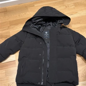 Svart pufferjacka från Canada Goose - Svart pufferjacka från Canada Goose med huva och dragkedja framtill. Jackan har quiltad design, två sidofickor och långärmade ärmar. Insidan är fodrad med mjukt material och har en klassisk Canada Goose-logga. Perfekt för kalla dagar.