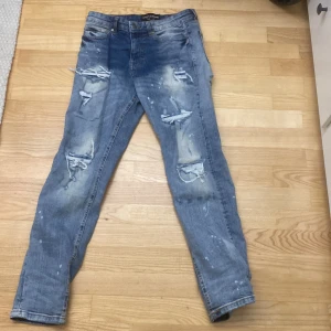 Supply & Demand ljusblå slitna jeans XS - Ljusblå jeans från Supply & Demand med coola slitningar och färgstänk. Modellen är smal och har klassisk femficksdesign. Jeansen har en modern, streetinspirerad look och är tillverkade i bomull. Perfekta för dig som gillar en edgy stil.