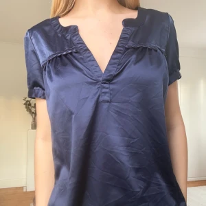 Mörkblå glansig blus med v-ringning - Mörkblå glansig blus med korta ärmar och v-ringad hals. Blusen har dekorativa broderade detaljer längs bröstet och lätt puffade ärmar. Materialet är slätt och har en satinlik finish. Köpt second hand men jag har bara använt den ett fåtal gånger så den är i bra skick.