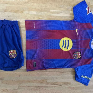 FC Barcelona Lamine Yamal matchtröja - Säljer en FC Barcelona fotbollströja med Lamine Yamal och nummer 10 på ryggen. Tröjan är kortärmad, har klassiska blå och röda ränder, gula detaljer och klubbmärke på bröstet. Materialet är lätt och andas, perfekt för match eller träning. Shorts medföljer med FCB-logga.