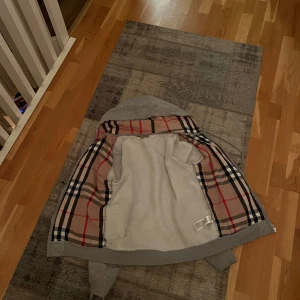 Grå hoodiejacka från Burberry - Grå hoodiejacka från Burberry med dragkedja och klassiskt rutigt foder i beige, svart och rött. Hoodien har huva med snörning, två fickor fram och är tillverkad i mjuk bomull. Perfekt för en chill och stilren look.
