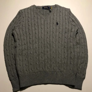Grå stickad tröja från Polo Ralph Lauren - Kabelstickad grå tröja från Polo Ralph Lauren med v-ringning och klassisk broderad logga på bröstet. Tröjan har ribbade muddar och är långärmad, perfekt för lager-på-lager under kyliga dagar.