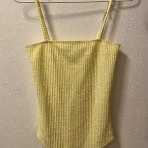 Gul ribbad bodysuit med smala axelband - Säljer en ljusgul bodysuit med smala axelband och ribbad struktur. Toppen har en rak halsringning och är tillverkad i ett mjukt, stretchigt material som sitter tight mot kroppen. 