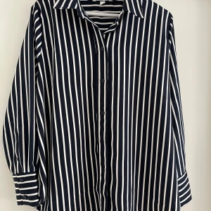 Randig skjorta från H&M, storlek L - Snygg skjorta från H&M i storlek L med vertikala marinblå och vita ränder. Klassisk krage, långa ärmar med manschett och knappar framtill. Skjortan har en loose passform och är tillverkad i mjuk polyester som känns skön mot huden.