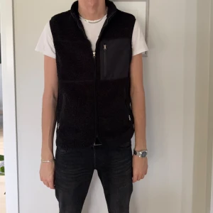Svart fleeceväst med dragkedja - Svart fleeceväst med hög krage och hel dragkedja framtill. Västen har en praktisk bröstficka med dragkedja i ett slätt material och två sidofickor med dragkedja. Perfekt för lager-på-lager och chill stil.