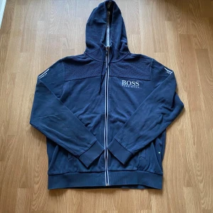 Blå hoodie från Hugo Boss - Snygg marinblå hoodie från Hugo Boss med dragkedja och huva. Tröjan har logga på bröstet, ribbade muddar och coola detaljer på ärmarna. Köptes för 1400kr. Säljer eftersom den inte får plats i garderoben:(