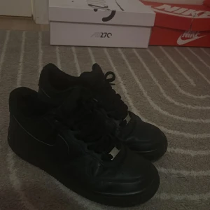 Svarta Nike Air Force 1 sneakers - Säljer ett par helsvarta Nike Air Force 1 sneakers i klassisk low-modell. Skorna har en stilren design med svart skinn, svarta snören och metallbricka med 'AF1' framtill. Perfekta för dig som gillar en clean och tidlös look.