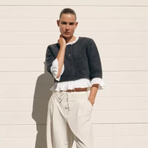 Grå kofta med volang från Zara - Supersöt grå kofta från Zara med vita volangdetaljer vid hals och nederkant. Koftan har två knappar framtill, är croppad och har trekvartsärm. Perfekt för dig som vill ha en unik och trendig look.