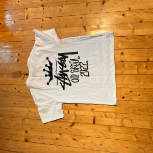 Vit Stüssy Old Skool 2022 t-shirt - Vit t-shirt från Stüssy med svart grafiskt tryck på både bröstet och ryggen där det står 'Old Skool 2022' och Stüssys ikoniska logga med krona. Klassisk passform och rund hals. Perfekt för dig som gillar streetwear och vill sticka ut med en clean look.