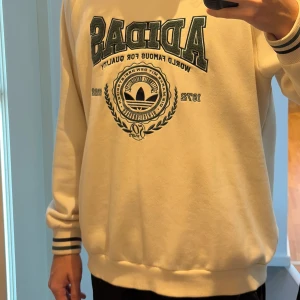 Beige Adidas sweatshirt med brodyr - Säljer en beige sweatshirt från Adidas med broderad logga och text framtill. Tröjan har ribbade muddar med svarta och gröna ränder samt rund halsringning. Perfekt för dig som gillar oversize stilren streetwear. Passar bra för personer som är 180-190!
