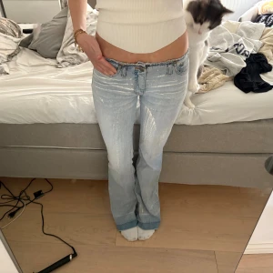Ljusblå bootcut jeans med rå kant - Säljer ett par ljusblå jeans med bootcut och låg midja. Jeansen har rå, fransig kant upptill och slitna detaljer framtill samt en kontrasterande bakficka. De är i klassisk denim och har en avslappnad, trendig vibe.