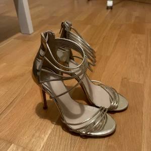  guldiga klackar - Säljer ett par klackar i metallic guld med öppen tå. Skorna har flera tunna remmar som går över foten och runt ankeln, vilket ger en cool och modern look. Dragkedja bak för enkel påtagning. Perfekta för att sticka ut med en glittrig vibe.