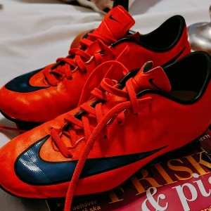 Nike Mercurial Fotbollsskor [stl. 38] - Säljer ett par Nike Mercurial fotbollsskor i stark orange med svarta och mörkblå detaljer. Skorna har snörning och platt sula med dobbar för gräsplan. Snygg, sportig design med Nike-logga på sidan och Mercurial-tryck på hälen. Perfekt för dig som vill sticka ut på planen.