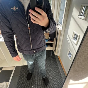 Mörkblå quiltad jacka från Morris - Säljer en mörkblå quiltad jacka från Morris Gent's i storlek medium. Jackan har snygga detaljer med broderad logga på bröstet, dragkedjor med läderdetaljer och klassisk krage. Tillverkad i 100% polyester och har lång ärm. Perfekt för höst och vår.