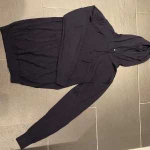 Marinblå hoodie från J.Lindeberg - Svart hoodie från J.Lindeberg i mjuk stickad bomull. Klassisk modell med huva och snörning, samt känguruficka framtill. Perfekt för en chill och stilren look. Långärmad och ribbade muddar vid ärmslut och nederkant.