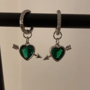 Örhängen med gröna hjärtan och pil silver  - Snygga örhängen i silverfärg med gnistrande stenar på ringen. Hängande berlocker i form av gröna hjärtan med pil igenom ger en cool och lekfull vibe. Perfekt för dig som vill sticka ut med ett unikt smycke med färg och detaljer.