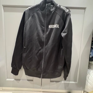 Svart track jacket från Hoodrich - Svart track jacket från Hoodrich med vit logga på bröstet och längs axlarna. Jackan har dragkedja framtill, ribbade muddar och ståkrage. Tillverkad i mjuk polyester för en sportig och clean look. Perfekt för dig som gillar streetwear.