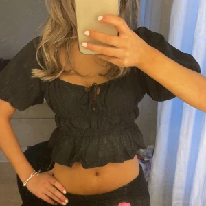 Svart croppad blus med puffärm - Säljer en svart croppad blus med korta puffärmar och volang nertill. Toppen har resår i midjan och vid bysten samt en söt knytning framtill. Perfekt för dig som gillar trendiga och feminina detaljer.