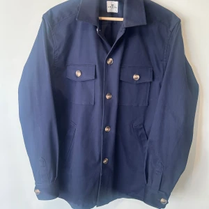 Mörkblå overshirt från John Henric - Stilren mörkblå overshirt från John Henric med klassisk krage, två bröstfickor med knappar och raka sidfickor. Jackan har långa ärmar och silverfärgade knappar framtill och vid ärmslut. Perfekt lager-på-lager-plagg för en clean look.