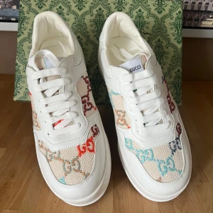 Gucci vita sneakers med färgglada GG-loggor - Snygga vita sneakers från Gucci med färgglada GG-loggor i rött, blått, orange och turkost på canvaspartier. Skorna har detaljer i vitt skinn, snörning och guldigt GG-tryck på hälen. Tjock sula med präglad GUCCI-text och GG-mönster undertill. Perfekt statement-piece för din outfit.