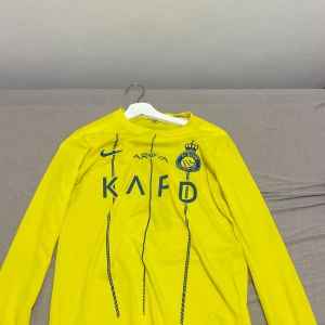 Gul långärmad Al Nassr Ronaldo tröja - Säljer en gul långärmad Al Nassr fotbollströja med Ronaldo och nummer 7 på ryggen. Tröjan har blå tryck och detaljer, klubbmärke på bröstet och Nike-logga. Tillverkad i lätt och ventilerande polyester, perfekt för match eller träning.