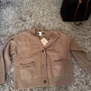 Beige kofta från Monki med knappar - Mysig beige kofta från Monki med två framfickor och ribbade detaljer vid ärmslut, nederkant och krage. Koftan har en avslappnad passform och stängs med två knappar framtill. Perfekt för lager-på-lager under höst och vinter.