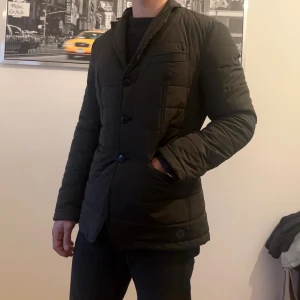 Mörkgrön quiltad kavajjacka - Stilren mörkgrön quiltad jacka med kavajkrage och knäppning framtill. Jackan har två fickor med lock och en bröstficka, samt diskreta sömmar som ger en snygg struktur. Perfekt för dig som vill ha en dressad look men ändå hålla dig varm.