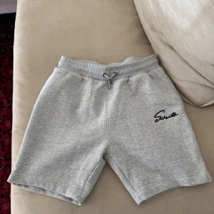 Grå sweatshorts med snörning - Säljer ett par grå sweatshorts från SikSilk med mjuk bomullskänsla och elastisk midja med snörning. Shortsen har en stilren design med logotyp broderad i svart på vänster ben. Perfekta för chill eller träning.