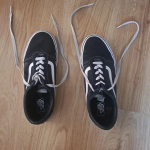 Svarta Vans Old Skool sneakers - Klassiska Vans Old Skool sneakers i svart canvas och mocka med vit sula och vit sidostrip. Skorna har vita snören och den ikoniska bruna gummisulan med rutmönster. Perfekta för en avslappnad streetstil.