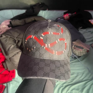 Gucci keps med orm och GG-mönster - Säljer en svart Gucci keps med klassiskt GG-monogram och en röd-vit orm broderad framtill. Kepsen har justerbart band bak med röd och blå rand. Tillverkad i bomull och polyester, riktigt snygg och unik accessoar för dig som vill sticka ut.
