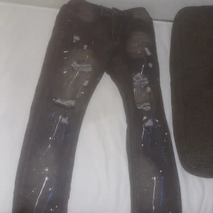 Svarta jeans med färgstänk - Säljer ett par svarta  jeans med coola färgstänk i vitt och blått samt slitna detaljer på framsidan. Jeansen har klassisk femficksdesign och är i stretchigt material för en tajt passform. Perfekta för dig som vill sticka ut med en unik streetstyle.