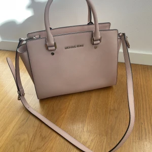 Ljusrosa handväska Michael Kors - Rosa handväska från Michael Kors med justerbar axelrem och silverfärgade detaljer. Väskan har ett rymligt huvudfack med dragkedja och plats för dina viktigaste ägodelar. Väskan är i riktigt fint skick, bortsett från en väldigt liten fläck på ena kortsidan. Annars är den i toppskick. Bustbag ingår.