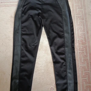 Svarta byxor med skinnpaneler - Snygga svarta byxor med coola skinnpaneler längs sidorna. Byxorna har en slim passform och midwaist, vilket ger en modern och stilren look. Perfekta för dig som vill ha något lite extra till outfiten.