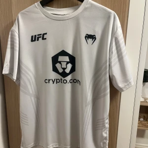 Vit UFC t-shirt från Venum - Säljer en vit UFC t-shirt från Venum med svart tryck och loggor på bröstet. Tröjan har korta ärmar och är gjord i ett lätt och svalt mesh material, perfekt för träning eller sportiga outfits. Snygga detaljer i tyget och modern passform.