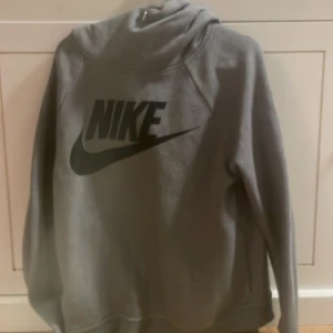 Grå Nike hoodie med svart logga - Säljer en grå hoodie från Nike med stor svart logga på bröstet. Tröjan har huva, långa ärmar och är tillverkad i mjukt material som passar perfekt för chill eller träning. Klassisk och enkel stil som funkar året runt.