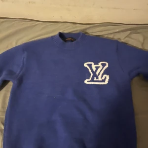 Blå stickad tröja Louis Vuitton - Säljer en blå stickad tröja från Louis Vuitton med stort vitt LV-logo på bröstet och ryggen. Tröjan har rund halsringning och långa ärmar, perfekt för dig som vill sticka ut med en lyxig och trendig look.