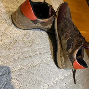 Valentino Rockrunners - Snygga Valentino Rockrunner sneakers med brunt och grönt camouflage-mönster, detaljer i vinrött och orange på hälen. Skorna har snörning, platt sula med nitar och är tillverkade i mocka, skinn och textil. Perfekt för dig som vill ha ett par varma, sköna och samtidigt stiliga skor nu i vinter. Pris kan diskuteras vid snabb affär, hör av dig om du är intresserad!😁
