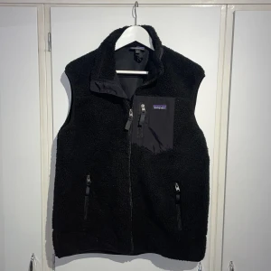 Svart fleeceväst från Patagonia - Svart fleeceväst från Patagonia med hög krage och hel dragkedja framtill. Västen har två sidofickor med dragkedja och en bröstficka med logotyp. Materialet är mjuk fleece och detaljer i syntet. Perfekt för lager-på-lager och outdoor. Priset är ej hugget i sten!✌🏽