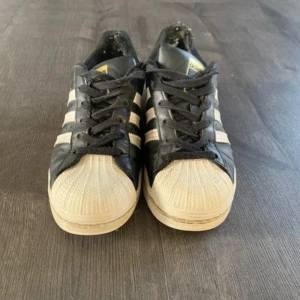 Adidas Superstar svarta sneakers strl 38 - Säljer ett par klassiska Adidas Superstar sneakers i svart och vitt med de ikoniska tre ränderna på sidan och snörning framtill. Skorna har rund tå och platt sula, tillverkade i skinn med detaljer i syntet. Adidas-logga på hälen och tungan. Perfekt för dig som gillar streetstyle.