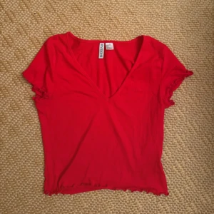Röd v-ringad topp  - Säljer en röd topp från H&M med v-ringning och korta ärmar. Toppen har volangdetaljer vid ärmslut och nederkant som ger en söt touch. Perfekt för dig som gillar enkel men snygg stil🫶