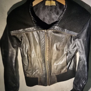 Svart skinnjacka Rock'n Blue - Svart skinnjacka från Rock'n Blue med ribbade detaljer på krage, axlar och nederkant. Jackan har en snygg dragkedja framtill och långärmade ärmar. Materialet är skinn och ribbad syntet, vilket ger en cool och modern look.