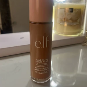 e.l.f. Halo Glow Liquid Filter - Glow booster i flytande form från e.l.f. i en varm brun nyans. Kommer i en cylinderformad flaska med ljusrosa lock och transparent botten. Perfekt för att ge huden en strålande finish och jämn ton.