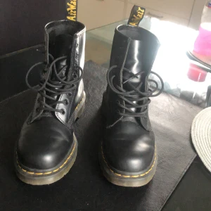 Svarta Dr. Martens boots strl 41 - Klassiska svarta Dr. Martens boots i skinn med rund tå, snörning och den ikoniska gula sömmen runt sulan. Grov platt sula och dragflik baktill med Dr. Martens-logga. Perfekta för dig som vill ha en edgy och tidlös stil. 