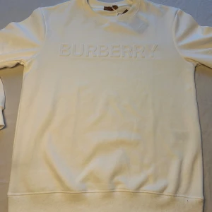 Vit sweatshirten från Burberry - Säljer en stilren vit sweatshirt  Burberry med broderad logga framtill. Tröjan har rund halsringning, långa ärmar och är tillverkad i mjuk bomull. Perfekt för dig som gillar klassisk och clean stil.