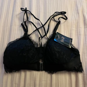 Svart spetsig korsett-bh - Svart korsett-topp från Hunkemöller med tunna axelband som korsas i ryggen. Stängs med silvrigt spänne framtill. Helt ny!